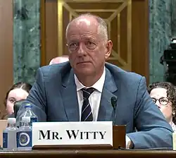 Andrew Witty, a bald, clean-shaven white man, wearing a light blue suit.