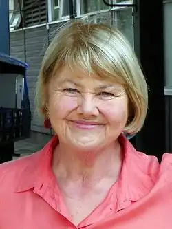 An image of a smiling woman. Her hair is primarily blonde, and she wears a pink shirt.