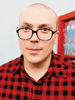 A man with black eyeglasses and red flannel looking at the screen, with a red cabinet and albums at the back