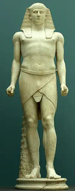 Marble statue of man wearing nemes headdress and pleated shendyt, standing with one leg forward and both fists clenched at his sides