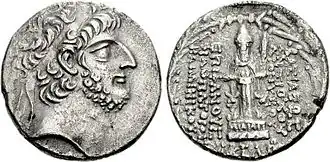Monochrome photograph of an ancient coin featuring a wreathed bearded male profile with curly hair on the left side. The right side depicts a standing figure with Greek inscriptions.