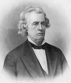 A man in his fifties, facing right, with chin-length, curly, white hair. He is wearing a white shirt, black tie, and black jacket.