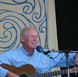 A white man with thinning white hair, sat at a microphone with an acoustic guitar, which he is tuning