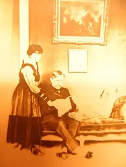 A man seated and a woman standing behind him in conversation in a richly furnished salon. The man reads a manuscript.