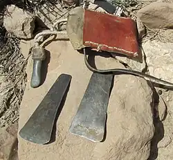 On a flat stone in the foreground, two metal blades or scrapers; in the background, another tool attached to a cord and a leather satchel.