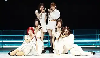 A group of six women performing onstage, while appearing in white streetwear clothing
