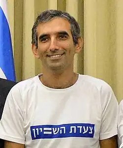 Photo of Dabush smiling at the camera. He wears a white t-shirt with Hebrew writing and has short, gray hair.