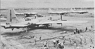 Four 4-engined World War II-era aircraft sitting on the ground at an airstrip. Groups of people are working near each aircraft.