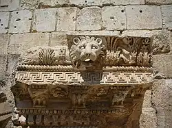 Photograph of an ancient stone carving of a lion's head surrounded by intricate patterns and foliage in front of a beige stone wall. The expression of the lion is fierce and detailed.