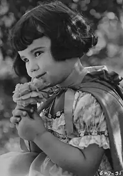 A young girl with short, dark hair wears a lace dress and cape, and holds a flower.