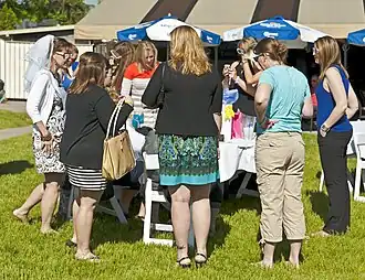 A group of women gathered around a table in an outdoor setting, dressed in a mix of casual and dressy styles. At the left of the image is a woman wearing a veil on the back of her head.