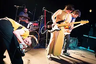Onstage at a small crowded venue, left to right: Katie Park playing guitar, Daoud Tyler-Ameen in the back playing drums, Emma Cleveland playing bass, and David Combs playing guitar