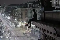 A person sits on the ledge of a building looking down at the street below