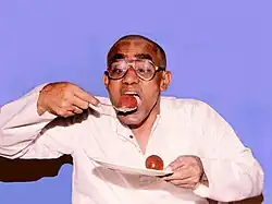 A man on a blue background is carrying a ledikeni (a redish brown ball about the size of a child's fist) in a spoon to his mouth with his right hand. His left hand holds a plate with another ledikeni on it. The ball visibly has some kind of light coloured syrup on them. there is a droplet of syrup on the spoon and the ledikeni on the plate is glistening.