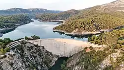 Aerial view of the Barrage de Bimont, a concrete gravity dam set in forested hills near Aix-en-Provence, with its curved structure retaining a large reservoir.
