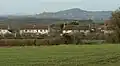 White painted houses with red roofs. In the background is a hill with a tower on it and in the foreground grass and hedgerows