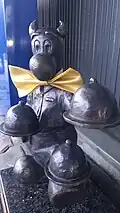 Bebok Maszket, ubrany z strój kelnera, niesie trzy przykryte tace. Dwie trzyma w rękach, trzecią balansuje na lewej stopie. Figurka przewiązana jest złotą kokardą