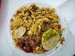 A bowl of several ingredients, including round crisps of dough, thin strands of dough, puffed rice, chutney, and vegetables