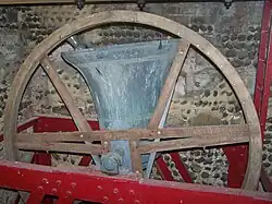 Large church bell named “Gabriel” mounted in a wooden wheel frame inside a stone tower. The bell is shown in the raised “up” position, ready for ringing, with red metal supports and flint-stone walls visible in the background.