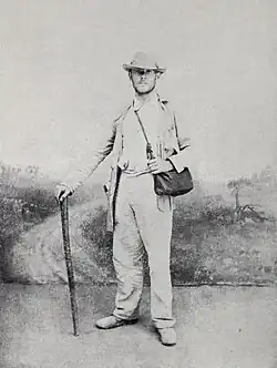 A young man with dark stubble in tan hiking clothes with brimmed hat, carrying a satchel and holding a walking stick