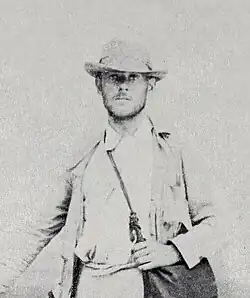 A faded black-and-white photograph of a young man with brimmed hat in a tan hiking outfit carrying a satchel