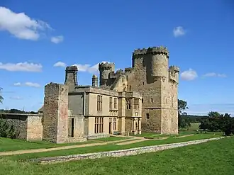 A large stone built ruined building. The nearer part is two stories with square windows; behind is a tall square keep with turrets and battlements. In the foreground is grass with a low stone wall; in the background a blue sky with a few white clouds.