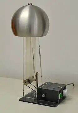 Large metal sphere supported on a clear plastic column, inside of which a rubber belt can be seen clearly: A smaller sphere is supported on a metal rod. Both are mounted to a base plate, on which is a small driving electric motor.
