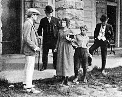 A vintage film still of a young woman holding books standing beside a boy and facing three men in suits and hats outside a stone building