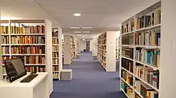 View down a long aisle in a library with tall bookshelves on both sides and a computer workstation on the left foreground.