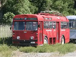 A red Wellington trolley bus with the trolleys tucked away parked up in a paddock by a fence. Bus sign says 'Karori Park number 12'.