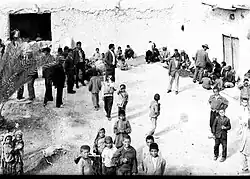 Black-and-white photograph of a wedding gathering in Bigherd, Iran, during the 1960s, with men, women, and children gathered in an outdoor courtyard beside whitewashed buildings.