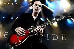 slightly blurry image of Billy Howerdel wearing dark clothing, playing a red electric guitar onstage, "Ashes Divide" written in large letters on the backdrop