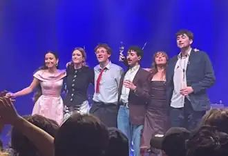 The six members of the band standing in a line on-stage, smiling with their arms on each other's shoulders.