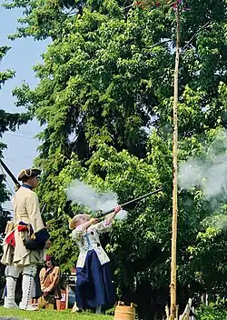 A women in a New France costume is shooting with a gun at a tree trunk to blacken it with gun powder