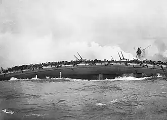 A sinking gray warship rolls over onto is side, its guns pointed skyward.