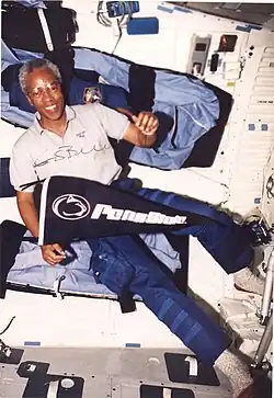 man wearing polo shirt and cargo pants, holding Penn State pennant, background interior of Space Shuttle