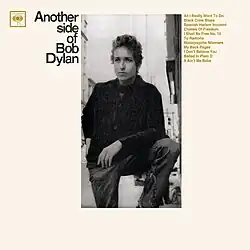 A black-and-white photograph of Bob Dylan standing with his foot raised, surrounded by thick white borders