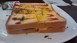 A sandwich with very thin vegetables between two slices of bread, topped with sev and ketchup and cut in four pieces.