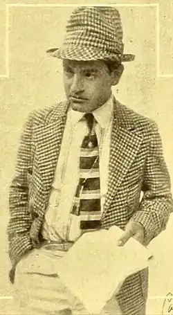 man wearing a checkered suit and matching hat holds a sheet of paper while looking to the side with a serious expression