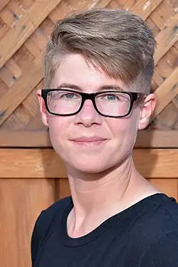 Woman's face with short light brown hair wearing black frame eyeglasses looking at camera smiling