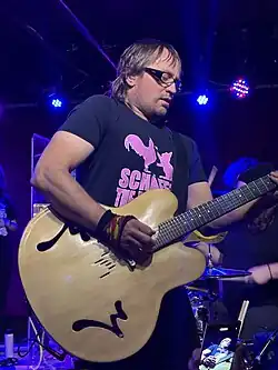 Wheatus frontman, Brendan B. Brown, wearing a black print t-shirt and playing an oversized acoustic guitar on a darkened stage; two other band members can vaguely be seen in background.