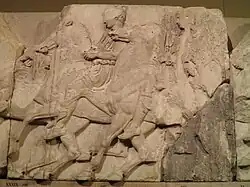 Slab of Greek sculpture showing cavalrymen riding: the lower right corner is coloured dark grey, while the rest is reasonably white in colour.