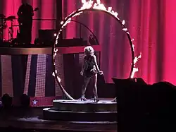 Image of a blond woman. She is standing in the middle of a fiery ring, wearing a black, white and gold outfit, with a pink led screen in front of her. A guitarist can be seen on her left.