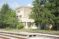 A three-story cream-colored building set among trees behind railway tracks in the foreground