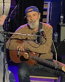 Bruce Sudano wearing a brown sweater, dark jeans, and a backwards blue cap, seated on a chair onstage in front of a microphone, holding an acoustic guitar, smiling directly at camera