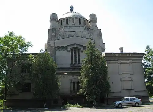 Weathered stone building with large dome, square towers, and symmetrical facade partially obscured by trees, with parked car in front