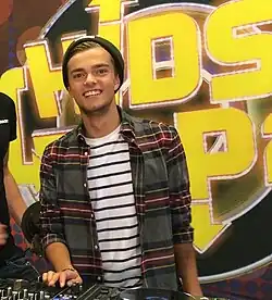 Photograph of a smiling teenager in front of the Kids Top 20 sign. He wears a beanie, a stripy white and black shirt and another open chequered shirt on top. His right hand rests on a dj table