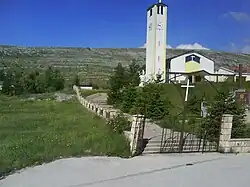 A photo of a white church on the outskirts of a village, taken from outside the front gate of the church, just outside of its garden. In the background is a stony hill.
