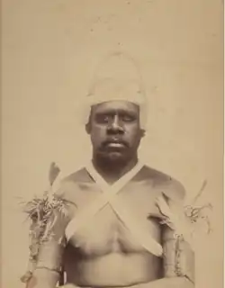 Portrait photograph of an Aboriginal man wearing a hat and decorative clothing made of rope and plants