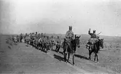 A column of around a dozen Foreign Legion troops on foot, followed by a similar number mounted on donkeys and led by two mounted officers/NCOs proceeding along a road